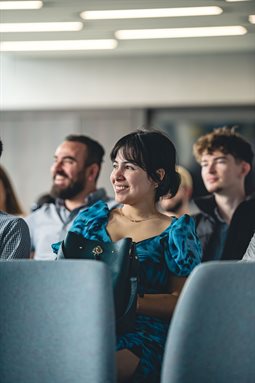 A person smiling they are sat down in an audience at a presentation