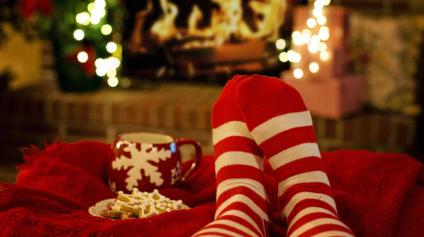 A pair of red and white striped socks on someone's feet with some snow flaked shaped cookies and a red mug with a white snowflake pattern