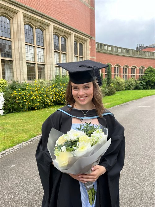 Female student dressed in a black graduation cap and gown holds a bouquet of flowers