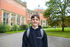 Male student with short hair stands in front of a red brick building, wearing a blue hoodie and grey backpack, smiling to the camera