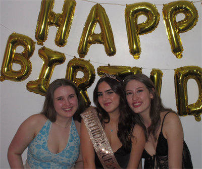 Three female students stand in front of gold balloons which spell 'Happy Birthday'. One of the female students is wearing a pink sash which says 'Birthday Queen'