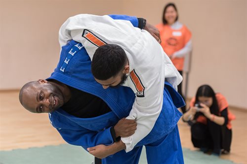 There are two male competitors in a grappling position. One male is wearing blue trousers and a black top with a blue jacket, is facing the camera. His opponent is wearing a white top and his face can't be seen. Behind them, slightly out of shot is a female photographer.