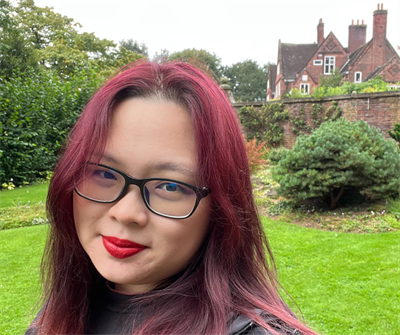 Female student with red hair and black glasses stands facing the camera. Behind her is a backdrop of grass, trees and a bricked building