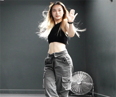Female student with blonde hair, wearing a black cropped top and grey cargo trousers posing with her palm facing in front of her. Behind her is a grey wall with a small silver fan in the corner.