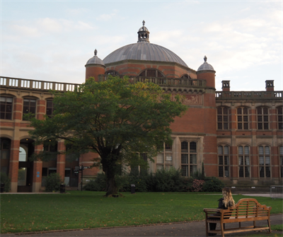 A female student sits on a brown wooden bench with her back towards the camera. She is facing a red brick building with a domed roof. There is also a tree with lots of green leaves in front of the building.