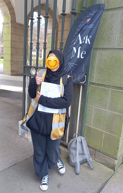 A student in a gold high visibility jacket stands by an 'Ask Me' sign with her thumbs up and an emoji covering her face.