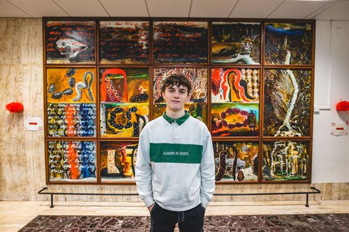 Male student wearing a grey jumper stands facing the camera with his hands in his pockets