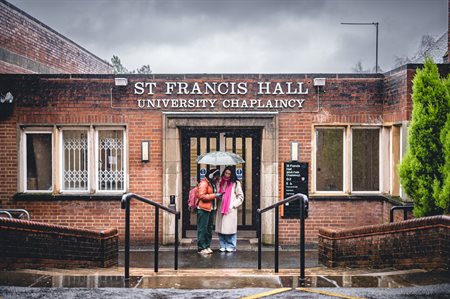 Two female students stand under an umbrella at the chaplaincy entrance.