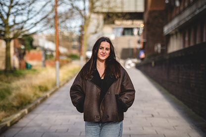 Female student with brown hair wearing a furry jacket standing on the pathway to Muirhead just by the Arts Building