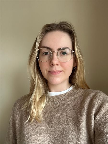 A young woman with blonde hair and glasses looks into the camera smiling. She is wearing a light brown jumper.