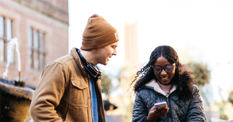 A female student is smiling looking at her mobile phone while a male student stands next to her