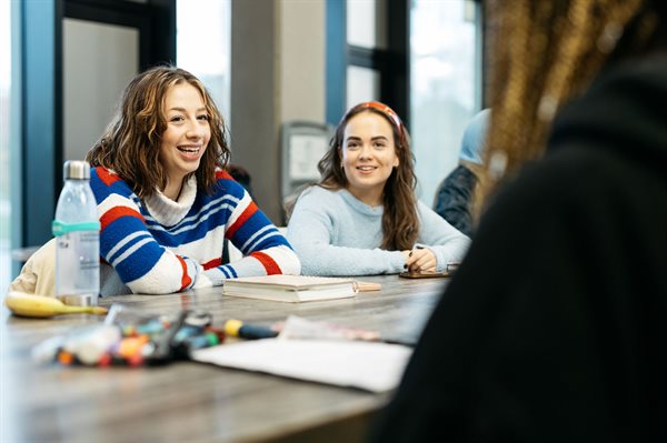 Two female students sit at a table smiling with their arms folded